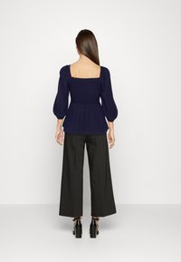 Top peplum blu navy con vita arricciata, maniche lunghe a sbuffo e scollatura quadrata, abbinato a pantaloni neri a gamba larga e tacchi neri spessi.