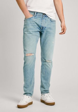 Light blue distressed jeans with rips, classic five-pocket design, made of denim, paired with white sneakers featuring brown soles.