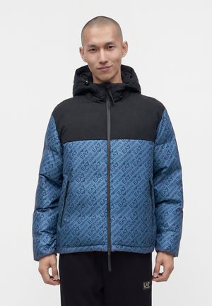 Homme portant une doudoune bleue et noire avec un motif répétitif du logo EA7 Emporio Armani, debout devant un fond blanc uni.