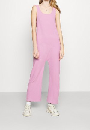 Kvinde iført en ærmeløs lyserød jumpsuit med vide ben, hvide sokker og hvide sneakers, stående mod en ensfarvet hvid baggrund.
