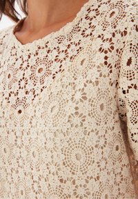 Gros plan sur un haut en dentelle beige crochetée avec des motifs circulaires et floraux porté sur une peau nue.