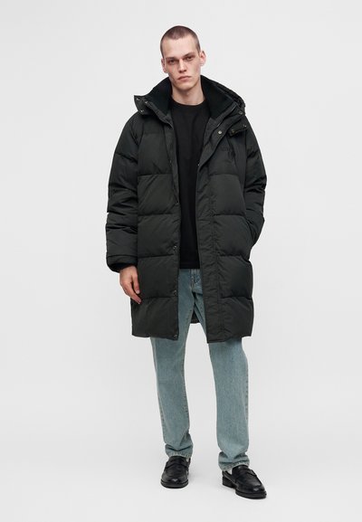 Jeune homme portant un long manteau matelassé noir, une chemise noire, un jean bleu clair et des chaussures habillées noires, debout devant un fond clair uni.