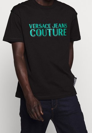 Mężczyzna w czarnej koszulce z krótkim rękawem z turkusowym tekstem "Versace Jeans Couture", w połączeniu z ciemnoniebieskimi jeansami na jednolitym tle.
