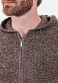 Sweat à capuche marron à fermeture éclair avec un tissu doux et texturé. Présente une fermeture éclair et une capuche confortable, affichant un design structuré mais décontracté.