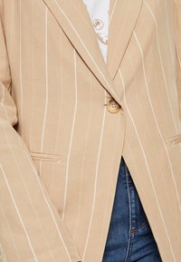 Blazer a strisce beige realizzato in tessuto leggero, con un bottone singolo, due tasche frontali e vestibilità sartoriale.