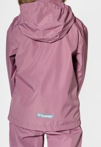 Lekka różowa wodoodporna kurtka z kapturem, elastycznymi mankietami i dolnym obszyciem. Na dole z tyłu znajduje się małe szare logo. Gładka, błyszcząca tekstura.