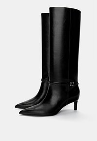Stivali in pelle nera al ginocchio con punte appuntite, tacchi sottili e una cinturino decorativo con fibbia attorno alla caviglia. Texture liscia, design elegante.