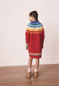 Červené pletené šaty s výrazným geometrickým vzorem v barvách duhy na ramenou, s dlouhými rukávy a žebrovaným lemem.