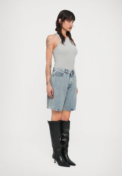 Γυναίκα με μακριά σκούρα μαλλιά που φοράει γκρι αμάνικο τοπ, φαρδιά τζιν σορτς, μαύρες ψηλές μπότες με τακούνι μέχρι το γόνατο, με εμφανή τατουάζ στον αριστερό της βραχίονα.