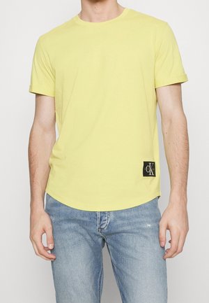 T-shirt jaune en coton avec un col rond et des manches courtes. Présente un écusson noir avec logo sur le bas à gauche.