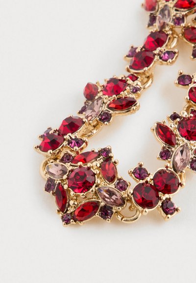 Klenot zlaté barvy s uspořádanými červenými, fialovými a bledě růžovými fasetovanými drahokamy různých tvarů a velikostí na bílém pozadí.