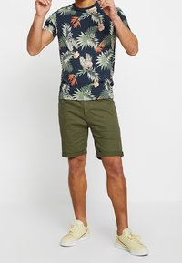 Camiseta de manga corta en color azul marino con un patrón de hojas tropicales y flores, combinada con pantalones cortos de oliva enrollados y zapatillas de color amarillo claro.