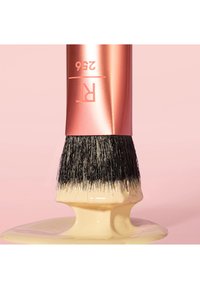 Pinceau de maquillage avec des poils noirs recouverts de fond de teint liquide beige sur un fond rose doux.