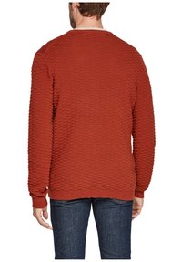 Czerwony sweter o fakturowanej powierzchni z prążkowanym wzorem, okrągłym dekoltem i długimi rękawami; w połączeniu z ciemnymi jeansami, o standardowym kroju.
