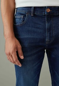 Jean en denim bleu foncé avec une coupe classique, cinq poches, fermeture à bouton en cuivre et détails de coutures jaunes contrastants.