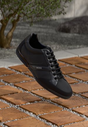 Schwarzer Leder- und Stoff-Sneaker mit Schnürsenkeln, "BOSS"-Logo an der Seite und auf der Zunge, dargestellt über einer Holzfliese und einem Kies-Außenbereich.