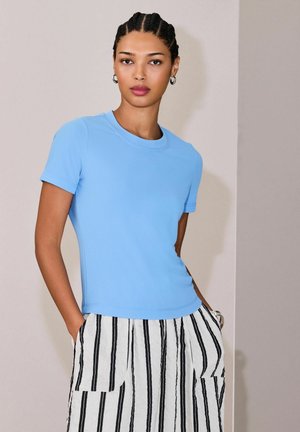 Punotut hiukset omaava nainen, joka pukeutunut vaaleansiniseen lyhythihaiseen paitaan ja valkoiseen hameeseen, jossa on mustia pystysuoria raitoja. Hän seisoo kädet taskuissa.