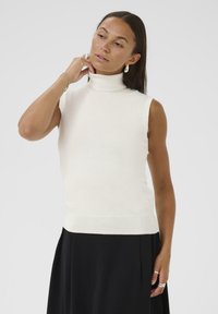 Kvinde med langt hår iført ærmeløs hvid turtle-neck og sort nederdel, der justerer sin ørering med den ene hånd nær sin hals.