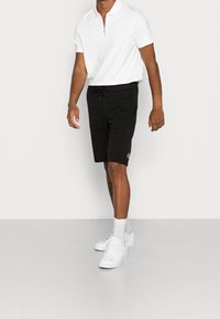 Pantalones cortos negros hasta la rodilla con una cintura elástica y cordón; combinados con una camiseta polo blanca con cremallera y un pequeño logo.