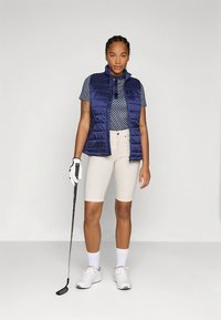 Colete acolchoado azul-marinho sobre uma polo azul estampada, combinado com calções bejes. Usado com ténis brancos e uma luva, segurando um taco de golfe.