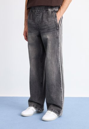 Person som bär löst sittande svarta jeans med sidostrek och vita sneakers, stående på en blå golv framför en vit vägg.