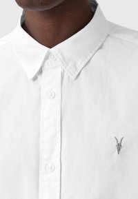 Camicia bianca in cotone con colletto button-down, caratterizzata da un piccolo teschio di capra ricamato sul petto sinistro. Tessuto liscio, design classico.