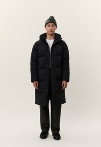 Jauns vīrietis, tērpts garā melnā pūkainā mētelī, tumšās biksēs, melnos apavos, baltā krekliņā un zaļā cepurē ar logotipu, stāvot pret vienkāršu fona virsmu.