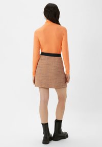 Langärmliges oranges Oberteil kombiniert mit einem karierten braun-orangenen Rock. Der Rock hat einen schwarzen Bund. Schwarze Knöchelstiefel runden das Outfit ab.