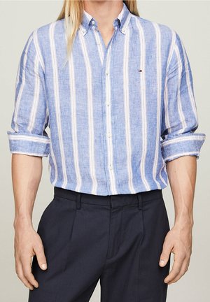 Man wearing a light blue and white vertically striped button-up shirt with sleeves rolled up and dark navy trousers.