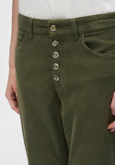 Pantalon vert olive avec cinq boutons métalliques ronds décoratifs et poches avant, porté avec un haut blanc, montrant la taille et le haut des jambes.