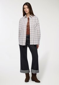 Camicia a quadri beige chiaro e marrone sopra una maglietta grafica marrone, abbinata a jeans larghi scuri arrotolati ai polsi e stivali marroni.