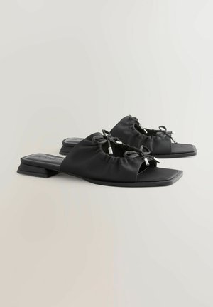 Sandali slingback neri con tomaia in tessuto testurizzato, caratterizzati da due fiocchi e dettagli metallici. Suola piatta e design a punta quadrata.