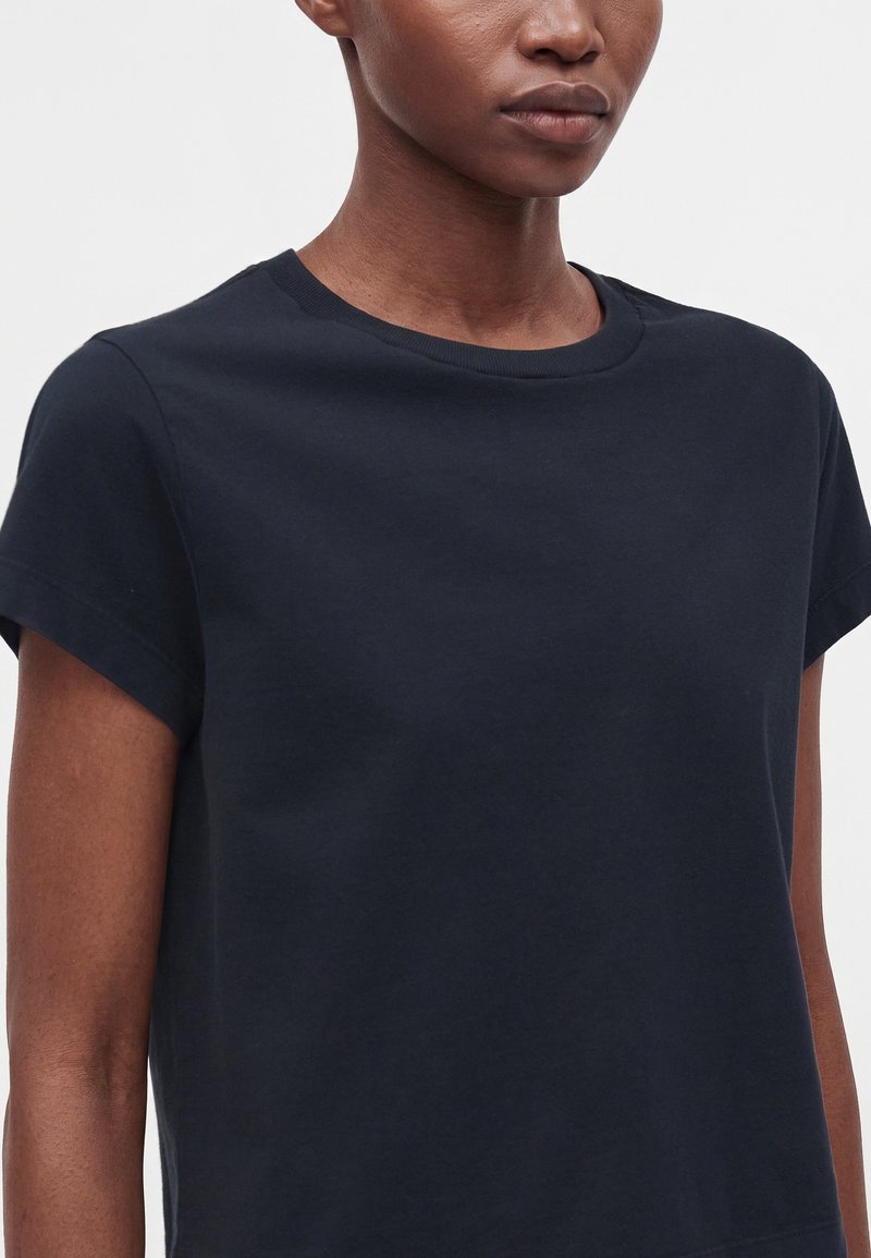 T-shirt noir à manches courtes et col rond porté par une personne à la peau foncée sur un fond blanc uni.