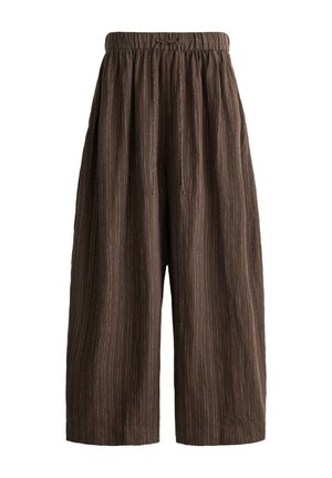 Persoană purtând pantaloni largi maro cu dungi subțiri, cu talie elastică și șnur, asortați cu o cămașă albă simplă cu mânecă scurtă.