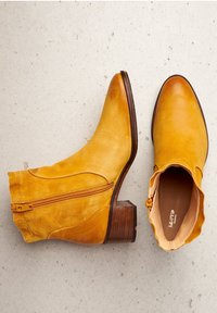 Žluté kožené kotníkové boty s špičatou špičkou, bočním zipem a vrstveným dřevěným podpatkem, vyznačující se hladkou texturou a minimalistickým designem.