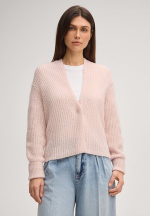 Frau mit langen braunen Haaren, die eine hellrosa gestrickte Strickjacke über einem weißen Hemd und weite hellblaue Jeans trägt, steht vor einem einfarbigen Hintergrund.