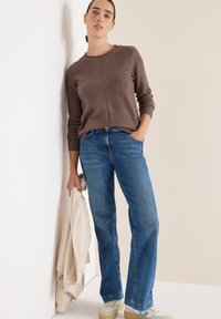Brauner Strickpullover mit rundem Ausschnitt, kombiniert mit hellblauen Jeans im geraden Schnitt. Graue und cremefarbene Kleidung in Hand gehalten. Legere Sneakers.