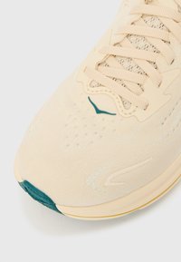 Αθλητικό παπούτσι ανοικτού μπεζ χρώματος με υφή στην επάνω επιφάνεια, τιρκουάζ λεπτομέρειες και επίπεδα κορδόνια. Διαθέτει στρογγυλεμένη μύτη και μαλακό πέλμα με αντίθετη άκρη.