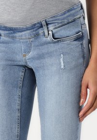 Jeans de mezclilla elásticos de color azul claro con un corte slim. Presentan un pequeño detalle desgastado en el muslo y bolsillos frontales estándar.