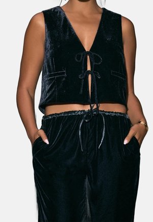 Femme portant un crop top sans manches en velours noir avec des liens à l’avant et un pantalon en velours assorti à cordon, mains dans les poches.