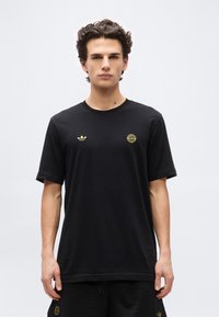 Νεαρός άνδρας στέκεται μπροστά, φορώντας μαύρο μπλουζάκι Adidas και μαύρο σορτς με χρυσά λογότυπα της FC Bayern Munich στο στήθος και στο πόδι.