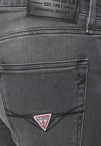Jeans de mezclilla gris oscuro con un acabado texturizado, que presentan una etiqueta de cuero negro y un parche triangular en rojo y blanco con el logotipo de "Guess" en el bolsillo trasero.