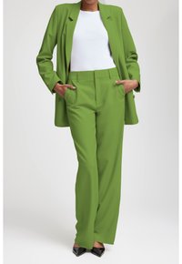 Kvinna i en grön kostym med blazer och vida byxor, vit topp, händerna i fickorna, svarta spetsskor.