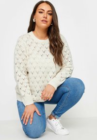 Cremefarbener Strickpullover mit einem Rautenmuster aus Spitze, kombiniert mit blauen Jeans und weißen Sneakers. Die Ärmel sind leicht lässig geschnitten.