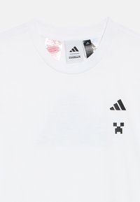 Valge T-särk, millel on mustad Adidas ja Minecraft Creeper logod ning nähtavad rõivamärgid krae sees vasakul rinnal.