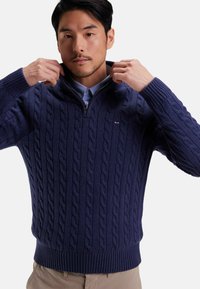 Pull en tricot bleu marine avec un col zippé, des poignets et un ourlet côtelés, fabriqué à partir d'un tissu en maille épaisse, doté d'un petit logo sur la poitrine.