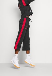 Sort sportswear-sæt bestående af en cropped jakke med røde striber, matchende bukser med røde side-striber og hvide sneakers med gummisåler.