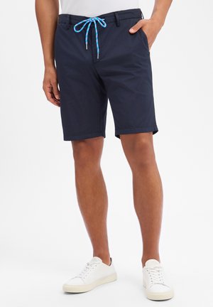 Mann trägt marineblaue Shorts mit Kordelzug, weiße Turnschuhe und ein weißes Hemd, steht mit einer Hand in der Tasche vor weißem Hintergrund.