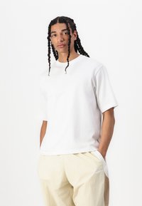 T-shirt branca de algodão com decote redondo e mangas curtas, combinada com calças bege claras com uma marcação subtil e uma textura suave.