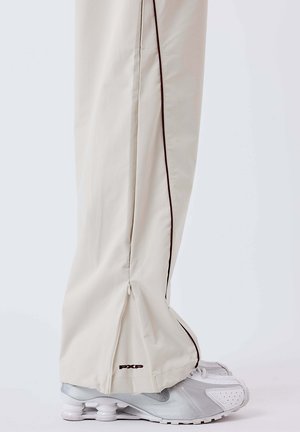 Pantalon de sport beige clair avec un détail de fermeture éclair latérale, présentant une bande contrastante marron foncé avec un liseré. Associé à des chaussures de sport argentées.